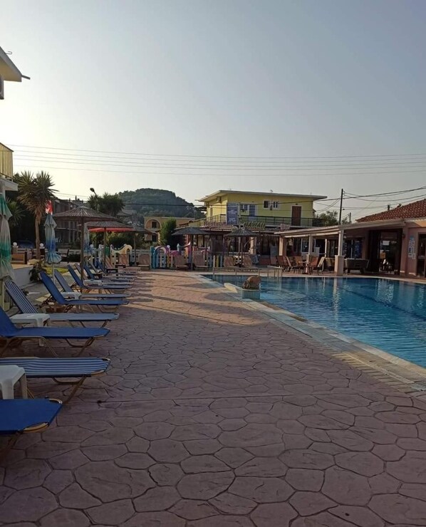 Zona de piscina con sillas de descanso y sombrillas, un restaurante y una colina al fondo.