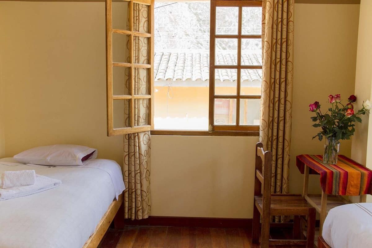 Una habitación con dos camas, una silla de madera, una mesita con un florero y una ventana con cortinas.