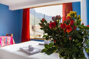 Habitación de hotel con cama, ventana con vista a la montaña y un ramo de rosas sobre la mesita de noche.