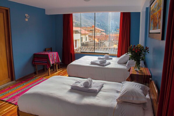 A hotel room with two beds, a window with a mountain view, a wooden floor, and a small table with a vase of flowers. - Tumy House B&B (Ollantaytambo)
