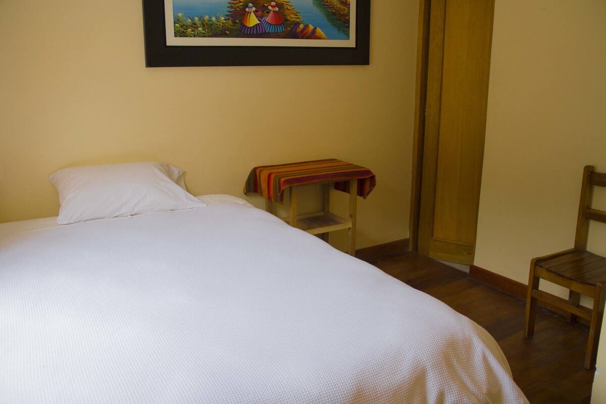 Una cama bien hecha con una colcha blanca, una mesita de noche de madera con un paño colorido y una silla de madera.