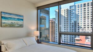 Una habitación de hotel con un ventanal grande, una cama, una mesita de noche, una lámpara y un cuadro en la pared.