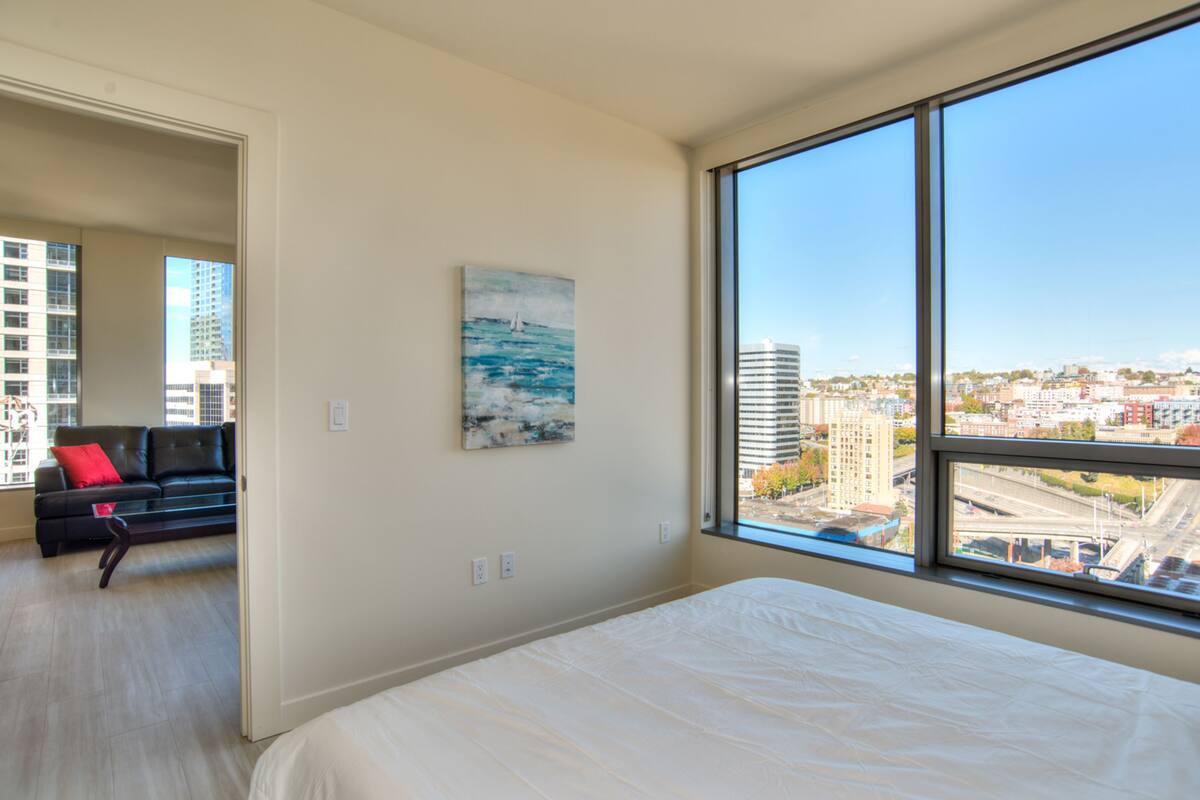 A modern hotel room with a large window offering a city view, a neatly made bed, a black leather sofa with a red cushion, and a painting on the wall.
