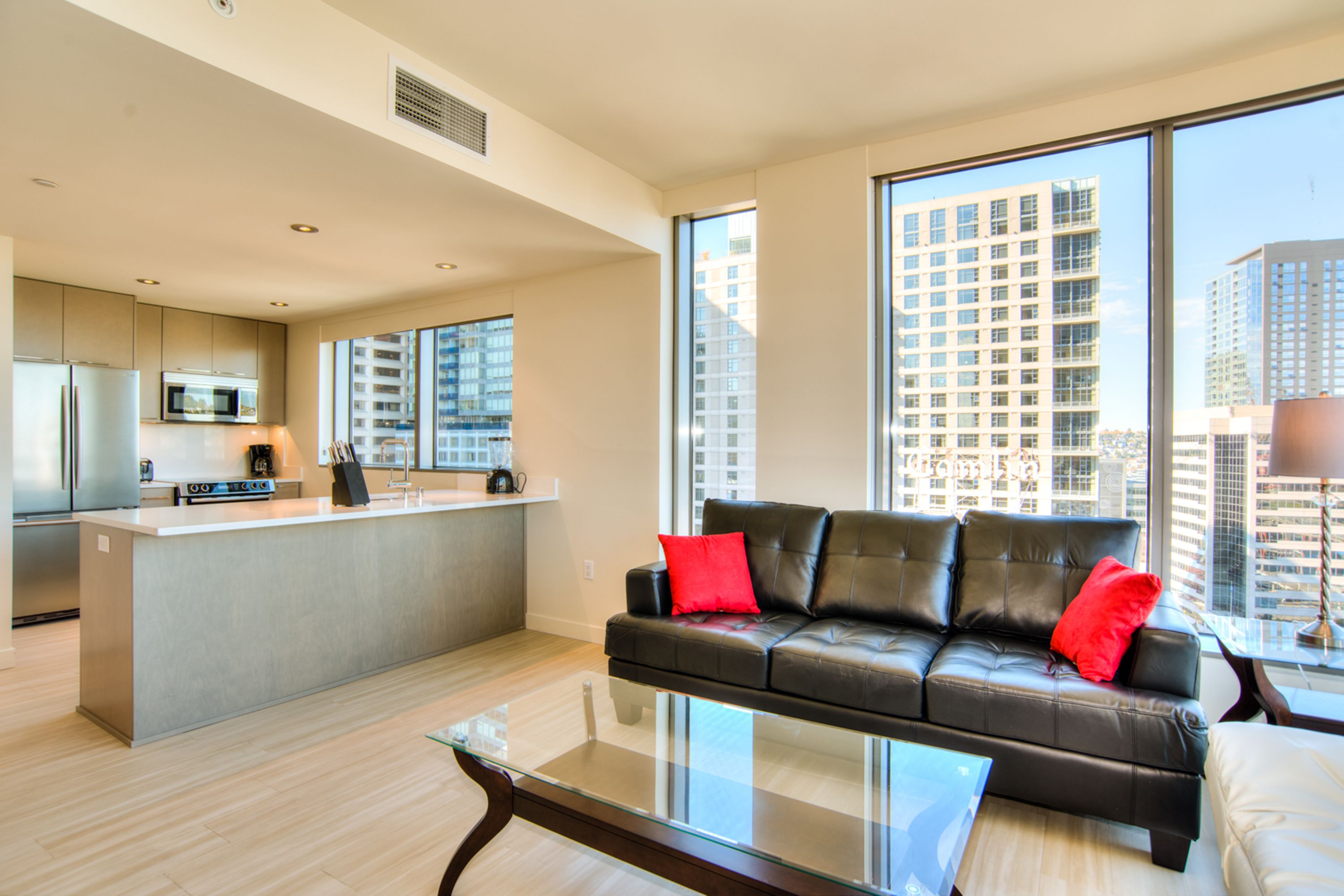 A modern living room with a black leather sofa, a glass coffee table, and a kitchen area with a microwave and stove.