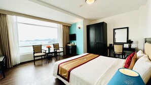 A hotel room with a large bed, a desk with two chairs, a TV, and a view of the sea.