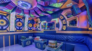 A vibrant, neon-lit bar area with blue seating and a karaoke machine.