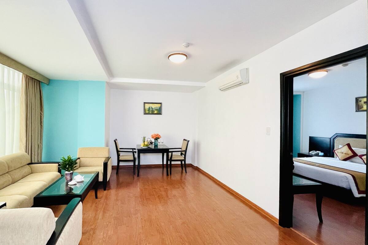 Habitació d'hotel amb sofà, taula de dinar amb cadires, taula petita amb una planta i llit amb capçalera.