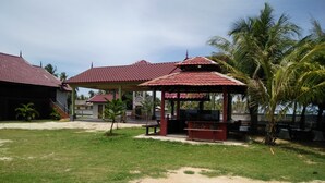 A covered outdoor area with a grill station, palm trees, and a building with a red roof. - Bayu Beach Penarek (Setiu)