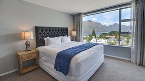 A bedroom with a large bed, two bedside lamps, a wooden nightstand, and a view of mountains through a large window. - Villa Two at Vailmont Queenstown (Queenstown)