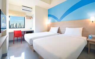Quarto de hotel com duas camas, uma escrivaninha, uma cadeira vermelha, vista para a cidade e um design de parede azul.