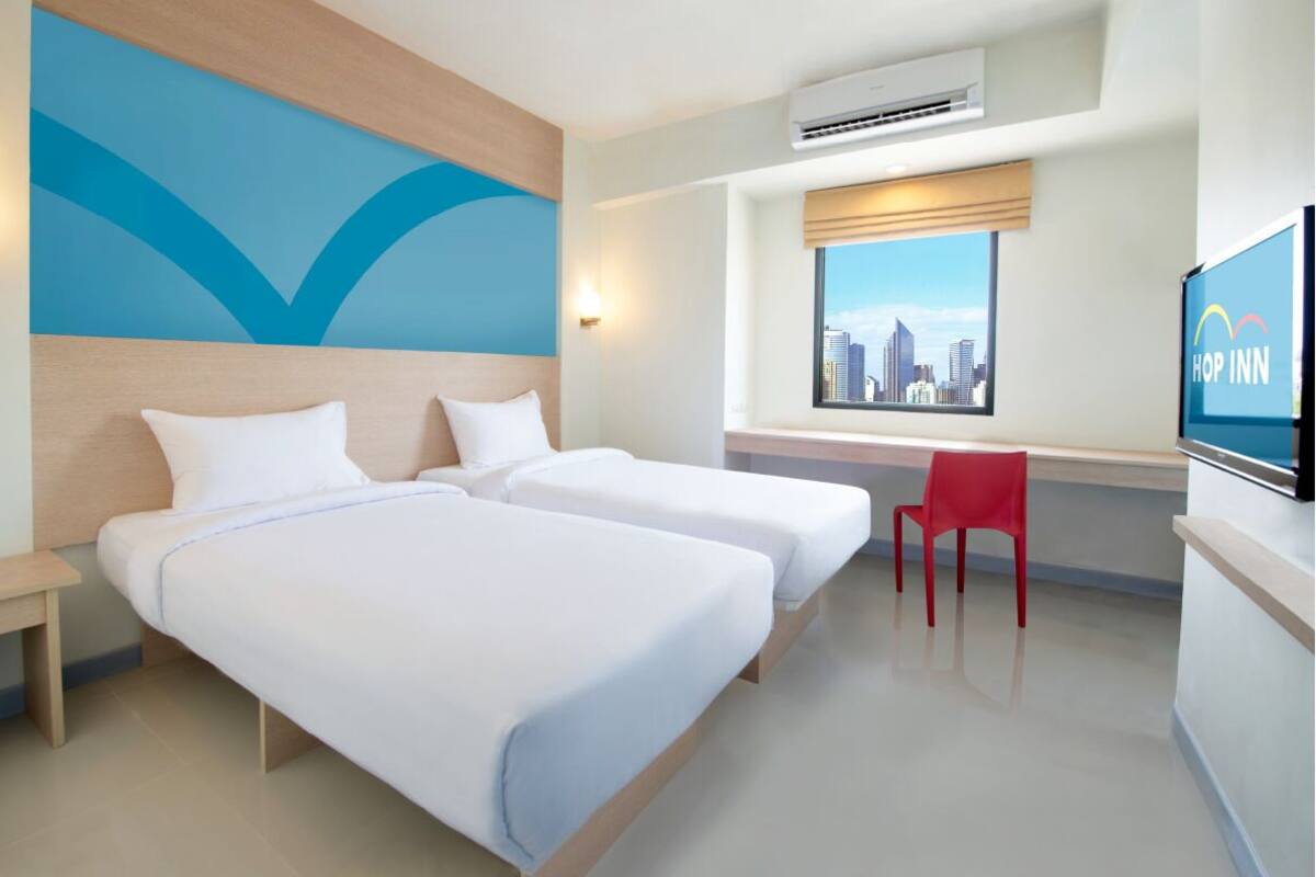A hotel room with two beds, a red chair, a city view, and a television displaying 'HOP INN'.
