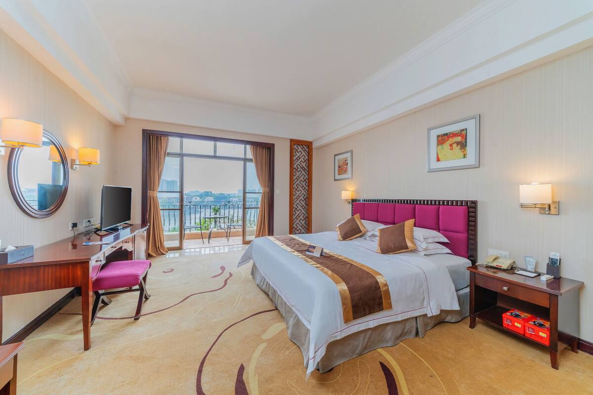 A hotel room with a large bed, a desk with a chair, a TV, and a view of the city through a large window.