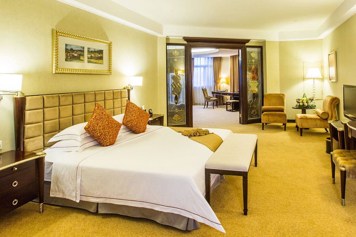 A hotel room with a large bed, a desk, a chair, a television, and a dining area visible through an open door.