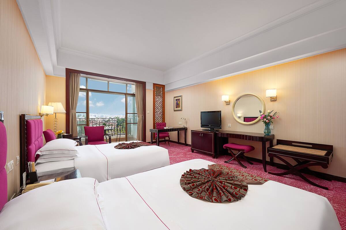A hotel room with two beds, a desk, a TV, and a large window offering a city view.