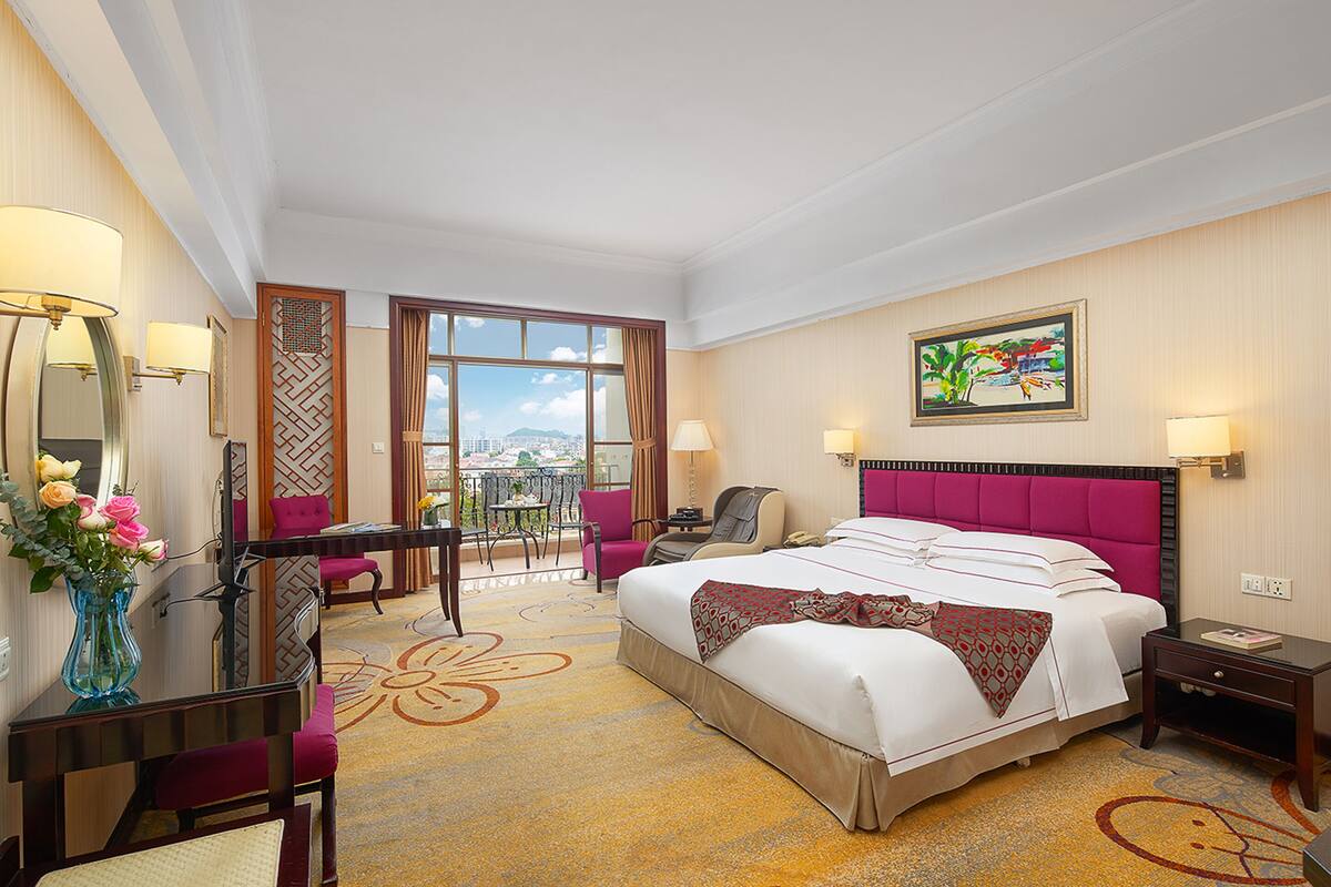 A hotel room with a large bed, a desk with a chair, a small table, a chair, a vase with flowers, and a window with a view.