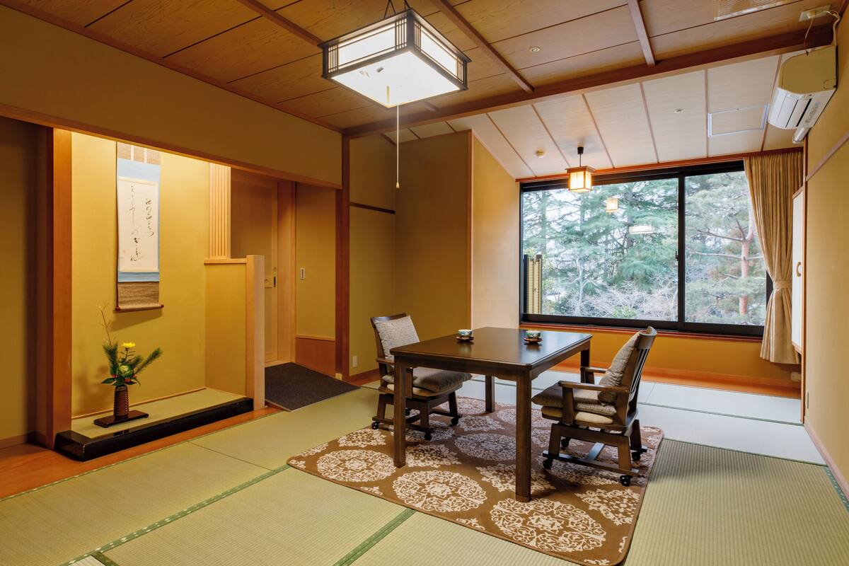 A traditional Japanese room with tatami flooring, a low table with chairs, a shoji screen door, and a view of trees through a large window.