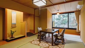 A traditional Japanese room with tatami flooring, a low table with chairs, a shoji screen door, and a view of trees through a large window.