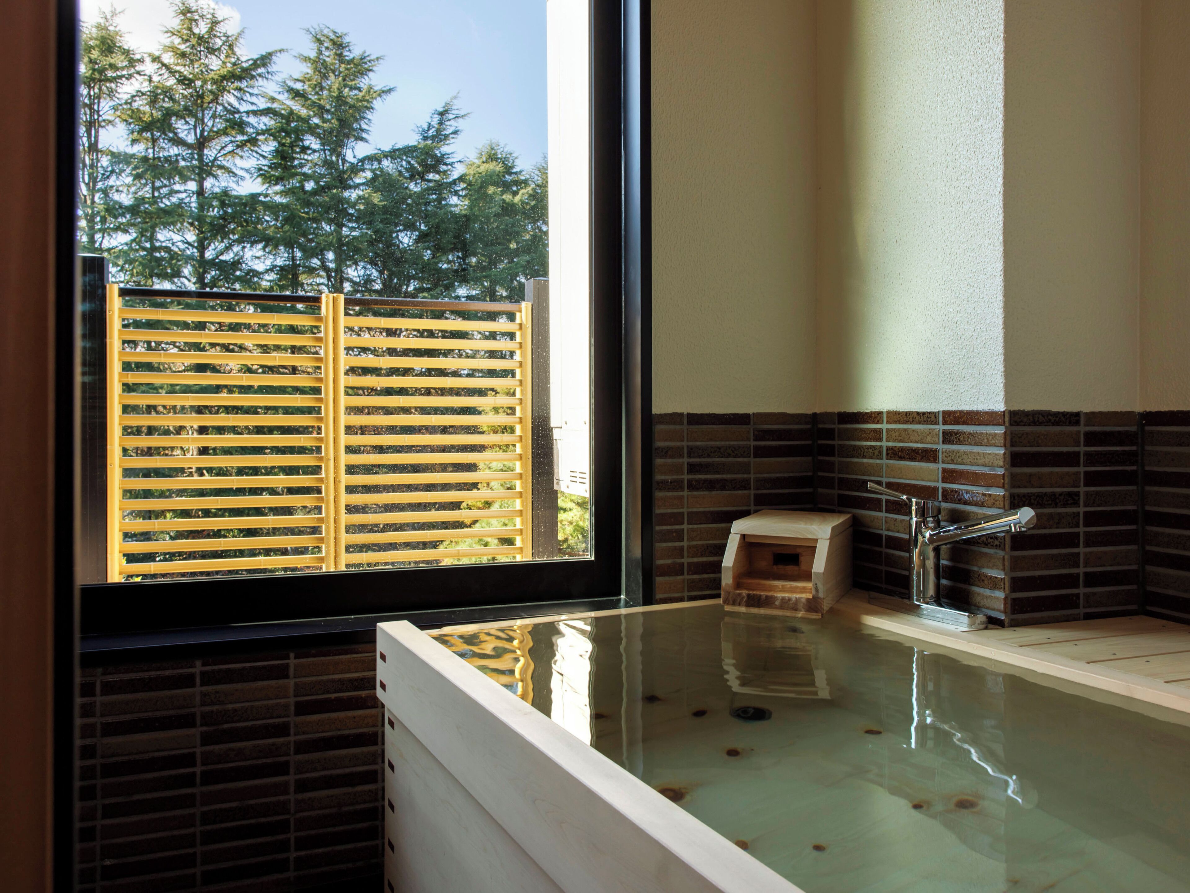 A modern bathroom with a glass-enclosed hot tub, a wooden bench, and a view of trees through a window with blinds.