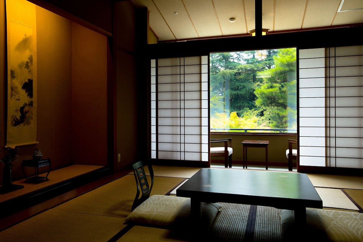 Ein traditioneller japanischer Raum mit Tatami-Böden, einem niedrigen Tisch und Schiebetüren mit Blick auf einen Wald.