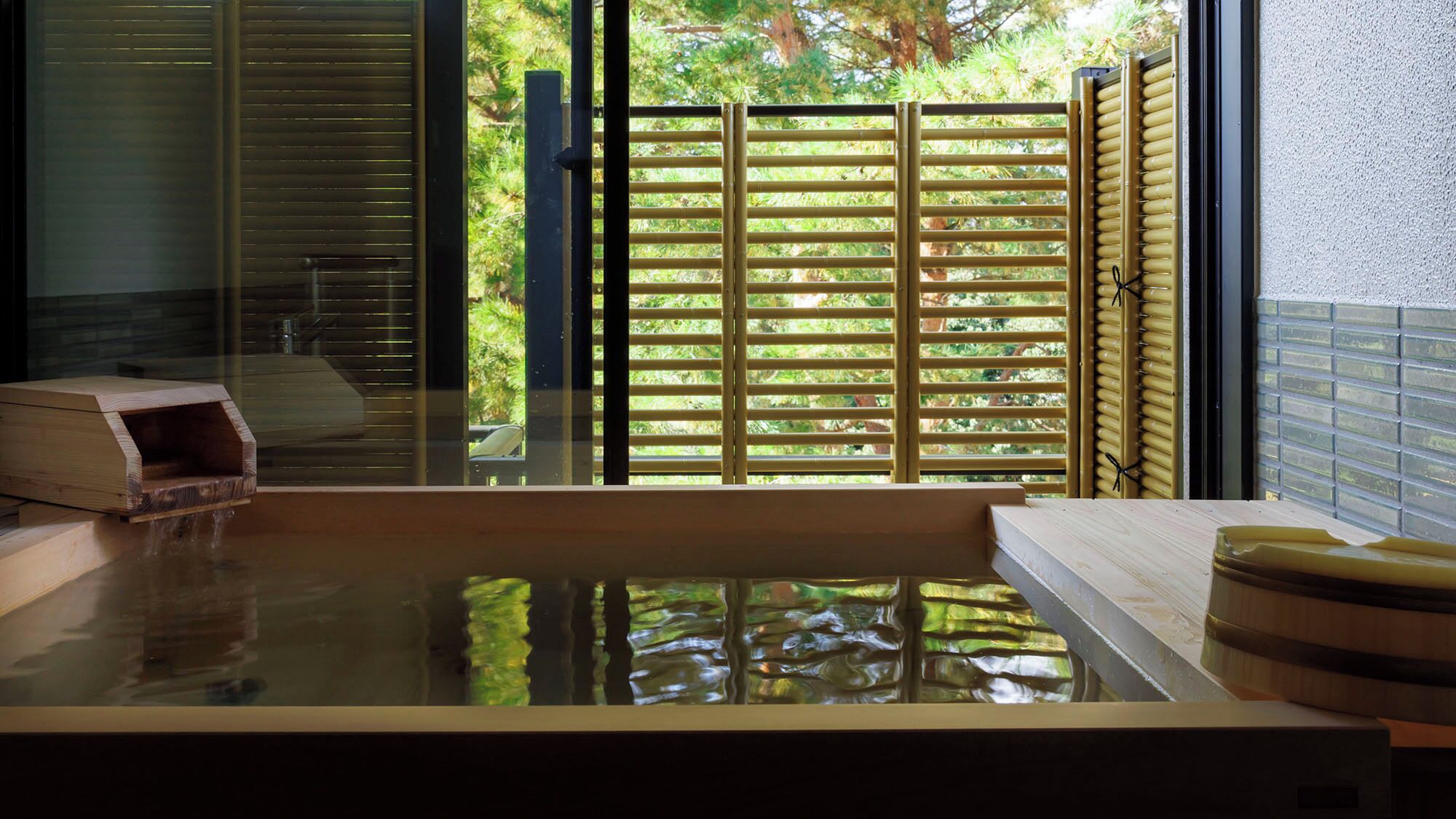 Un jacuzzi con un banco de madera, una estructura de madera de la que sale agua, y una valla de bambú al fondo.