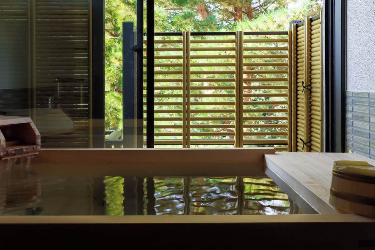 A hot tub with a wooden bench, a wooden structure with water flowing out, and a bamboo fence in the background.