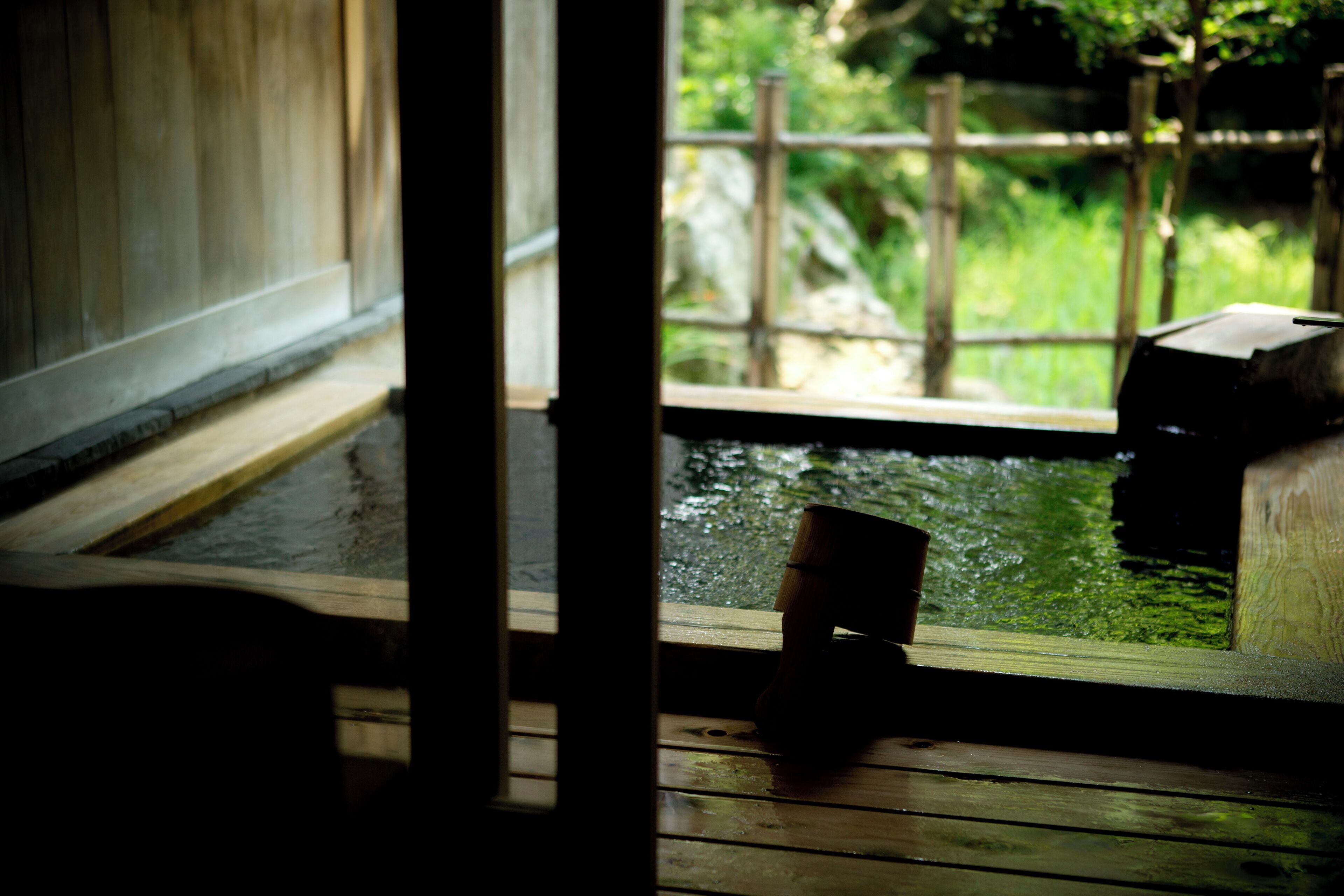 Una terraza de madera con un banco y una mesita, con vistas a un jardín con estanque y follaje.