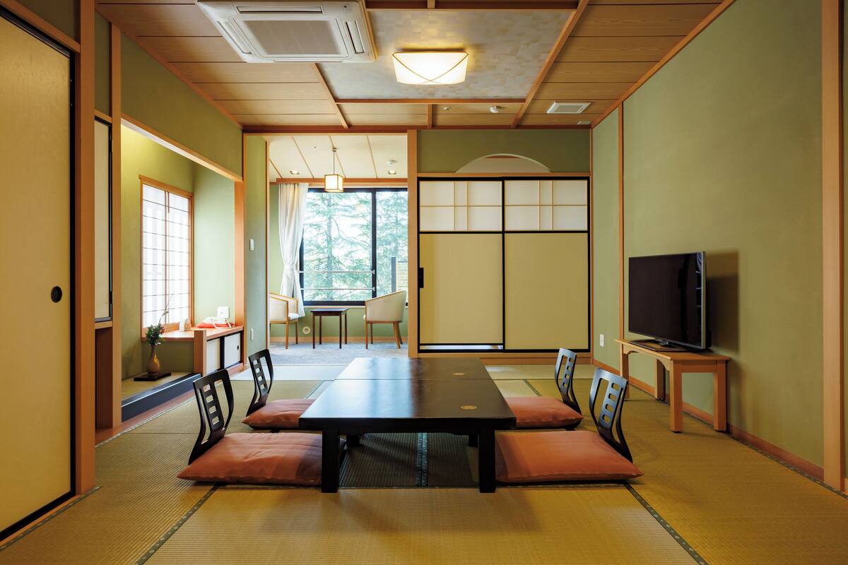 A traditional Japanese-style room with tatami flooring, a low table with cushions, a television on a stand, and sliding doors leading to another room.