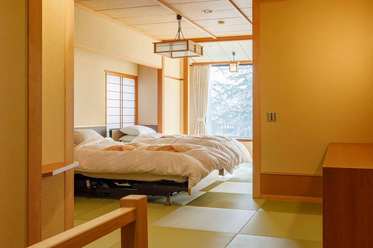 A traditional Japanese-style room with tatami flooring, a bed with white bedding, a wooden desk, and a sliding door.