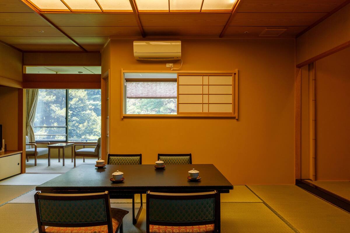 A traditional Japanese room with tatami flooring, a low table with two chairs, a window with shoji screens, and an air conditioning unit.