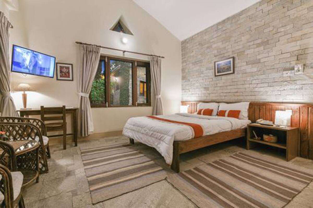 A bedroom with a bed, wooden headboard, a nightstand, a TV, a window with curtains, and a stone wall.