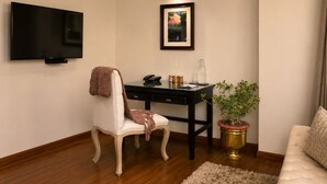 A living room with a flat-screen TV mounted on the wall, a desk with a telephone, a framed picture, a potted plant, and a sofa.