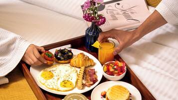A tray with breakfast items including eggs, croissants, and a beverage, placed on a bed with a magazine and a vase of flowers.