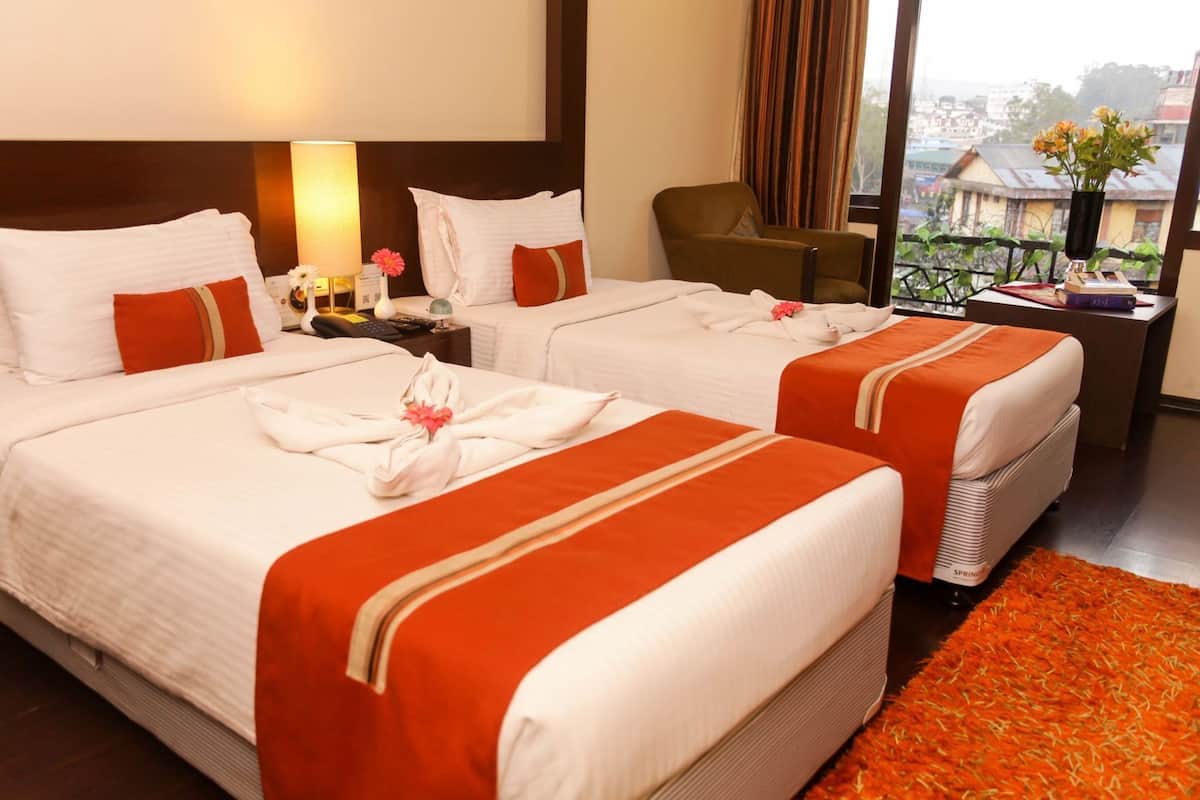 A hotel room with two beds, a nightstand with a lamp, a chair, and a view of the city.