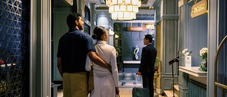 Un couloir d’hôtel avec un sol en marbre, un lustre et des panneaux indiquant les différents espaces, comme le « service de conciergerie » et la « réception ».