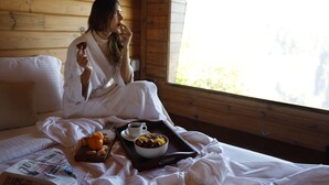 A person sitting on a bed in a wooden cabin room with a tray of food and a newspaper. -  Polo Cherrapunjee Resort  (Shillong)