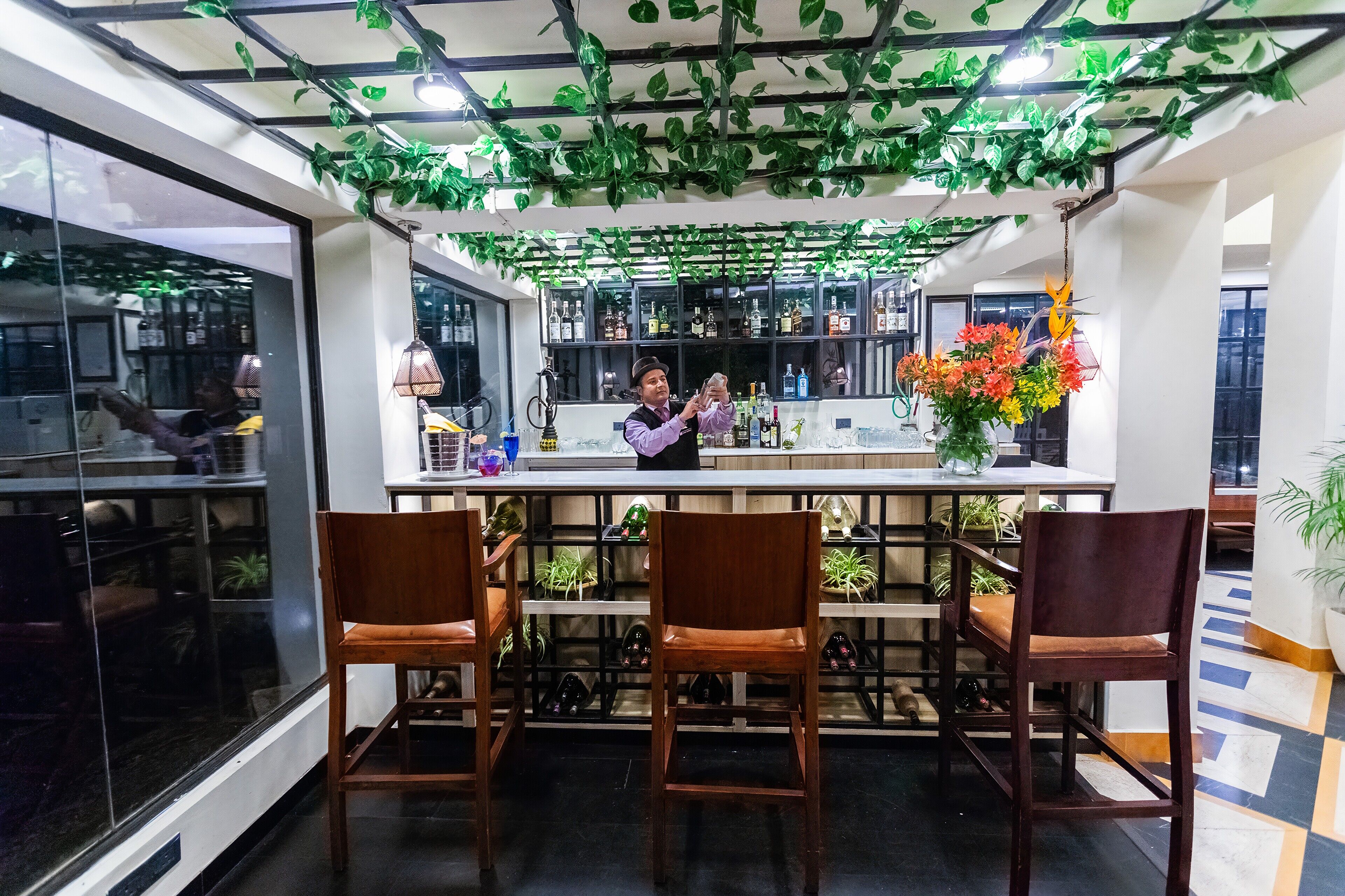 A bar area with wooden chairs, a bartender, and shelves with bottles.
