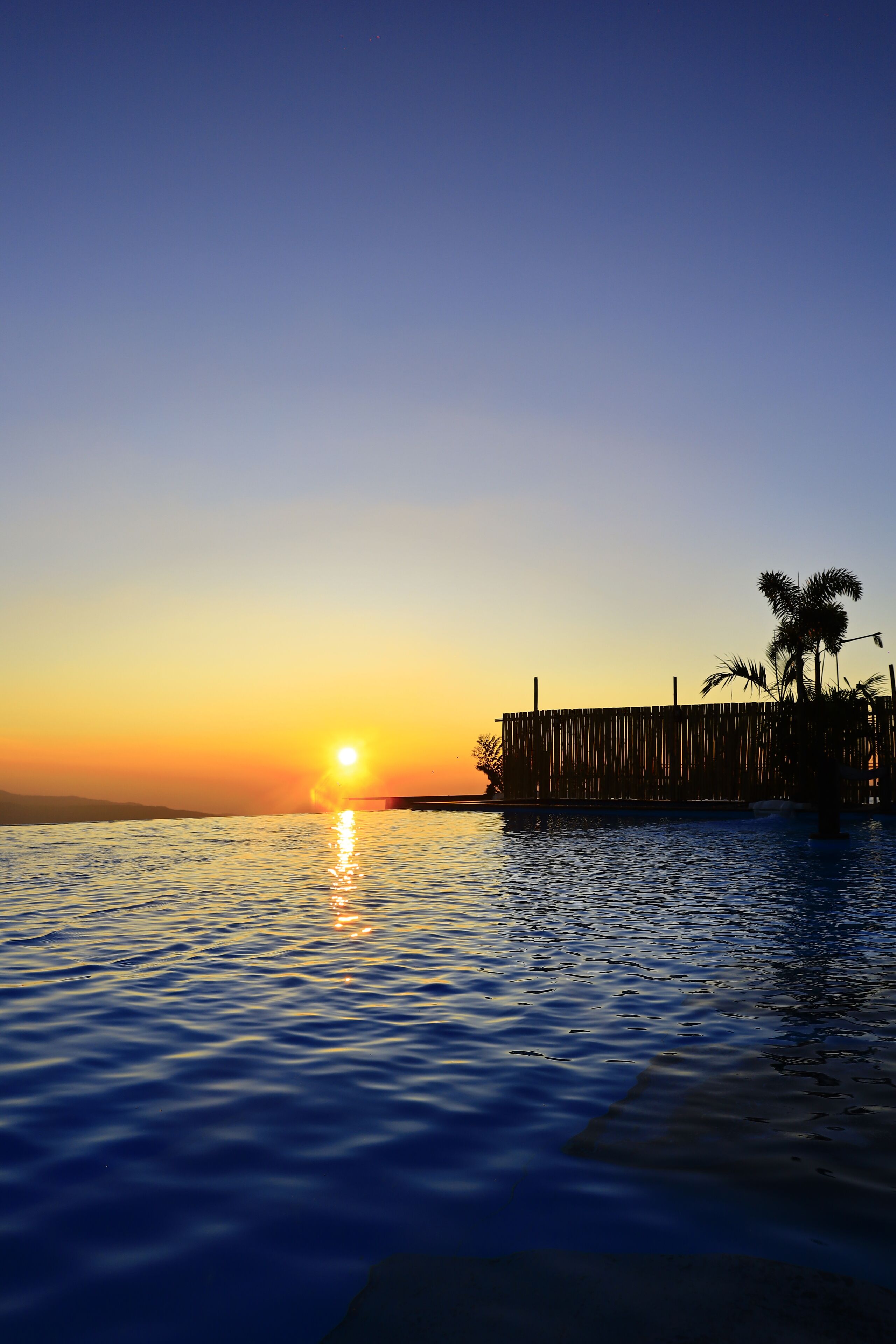 A sunset over a calm body of water with a wooden dock and palm trees.