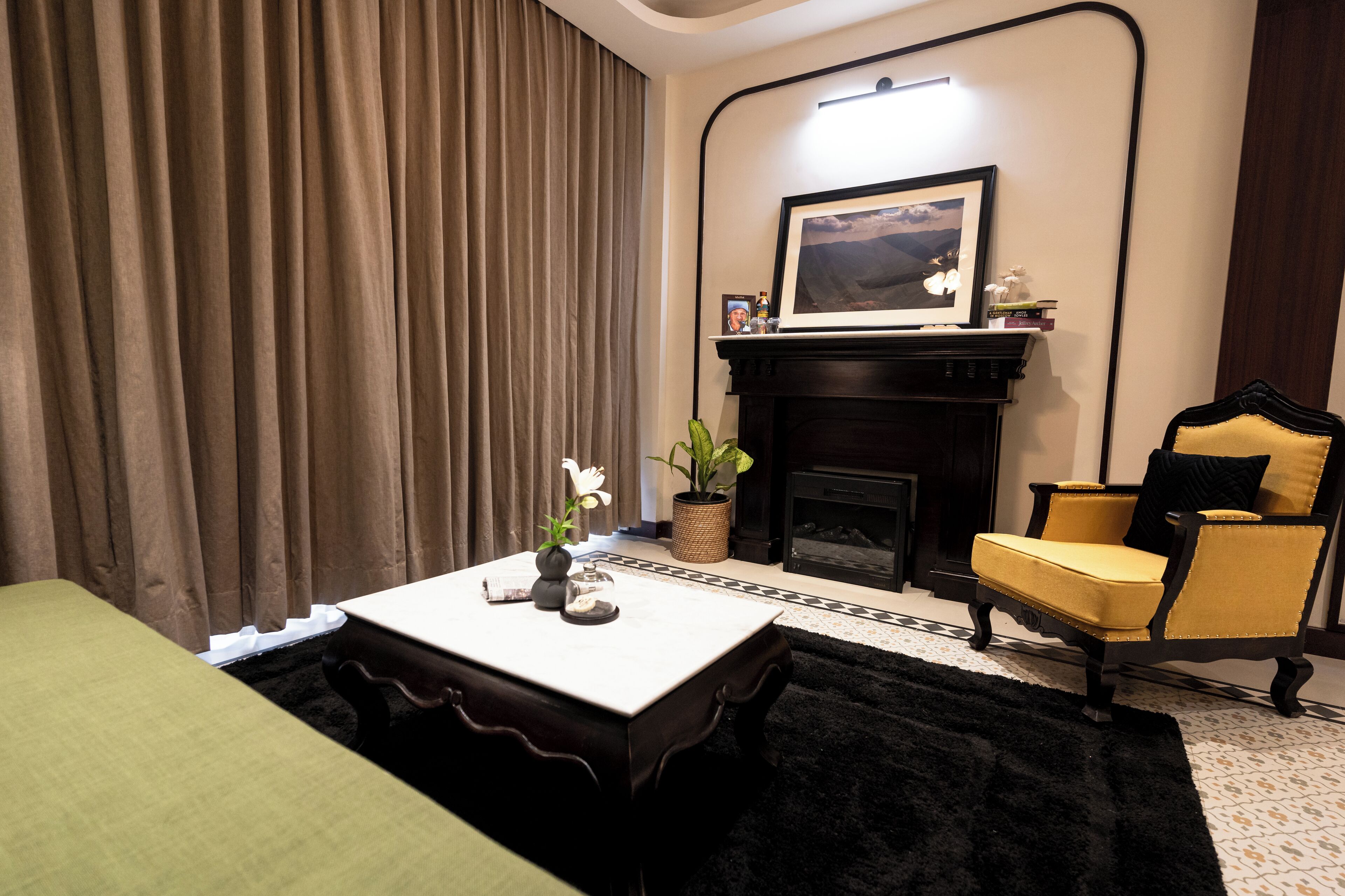 A hotel room with a bed, a table with a vase and a picture frame, a chair, a fireplace, and a large window with curtains.