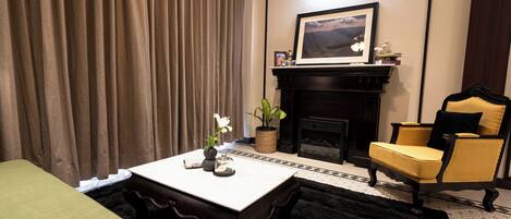 A hotel room with a bed, a table with a vase and a picture frame, a chair, a fireplace, and a large window with curtains.