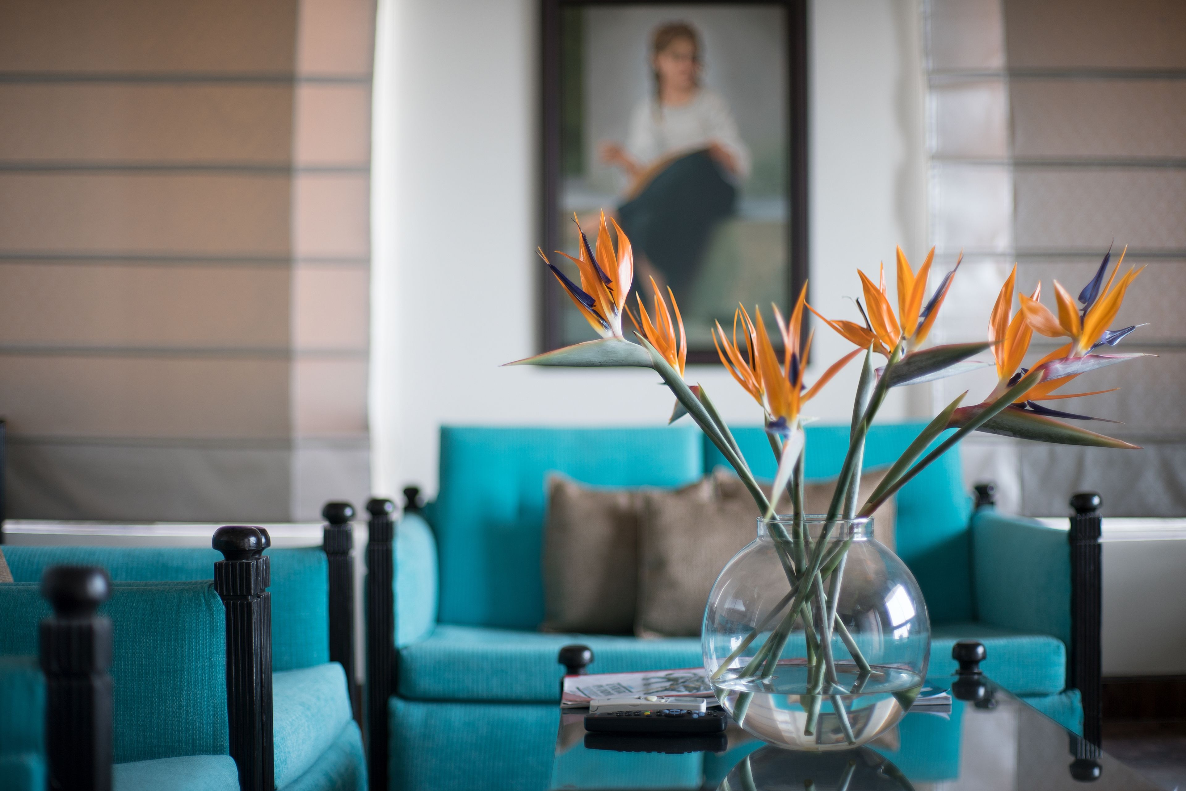 A living room with a teal sofa, a glass coffee table with a vase of bird of paradise flowers, and a framed picture on the wall.