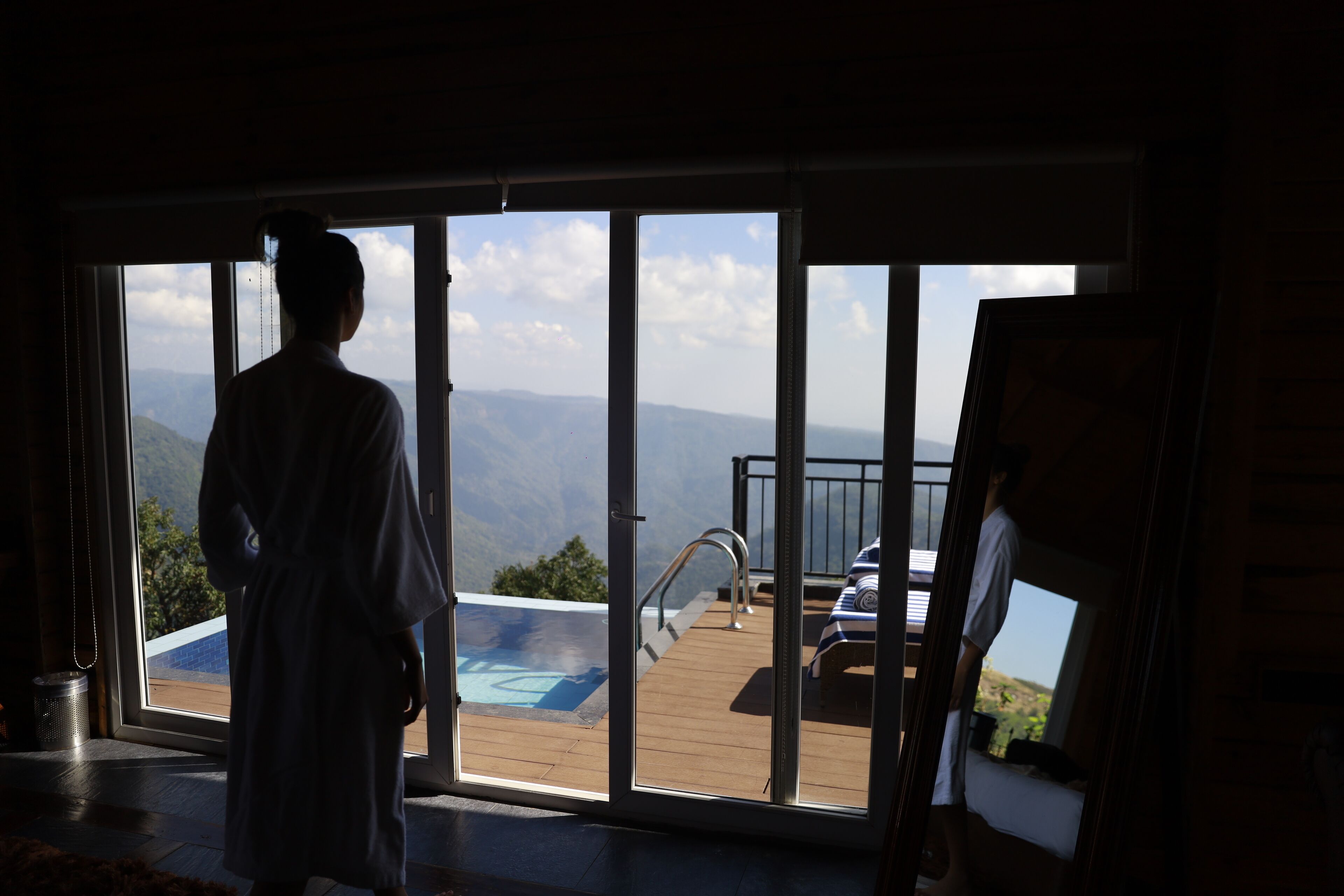 A person in a robe standing by a large sliding glass door leading to a deck with a pool and outdoor seating.
