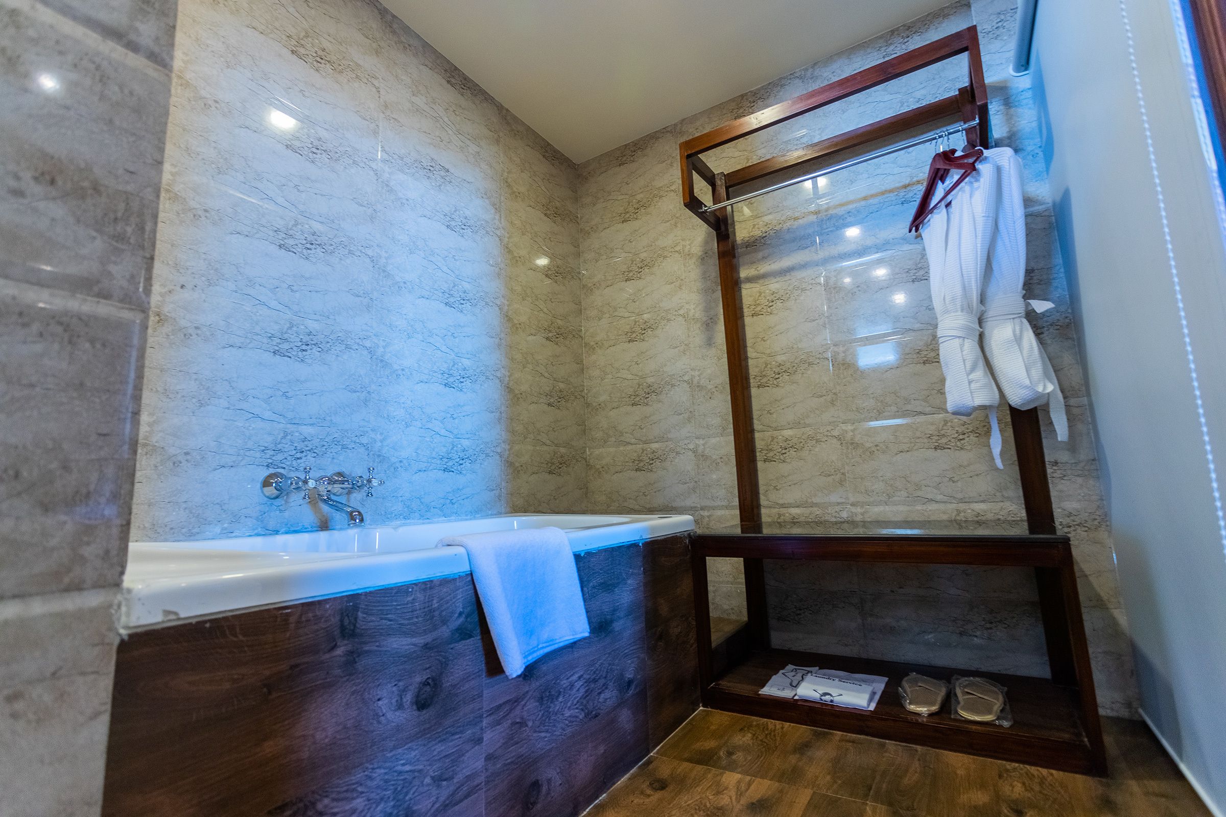 A modern bathroom with a marble wall, a wooden bathtub, a glass shower enclosure, and a wooden vanity with a towel rack.