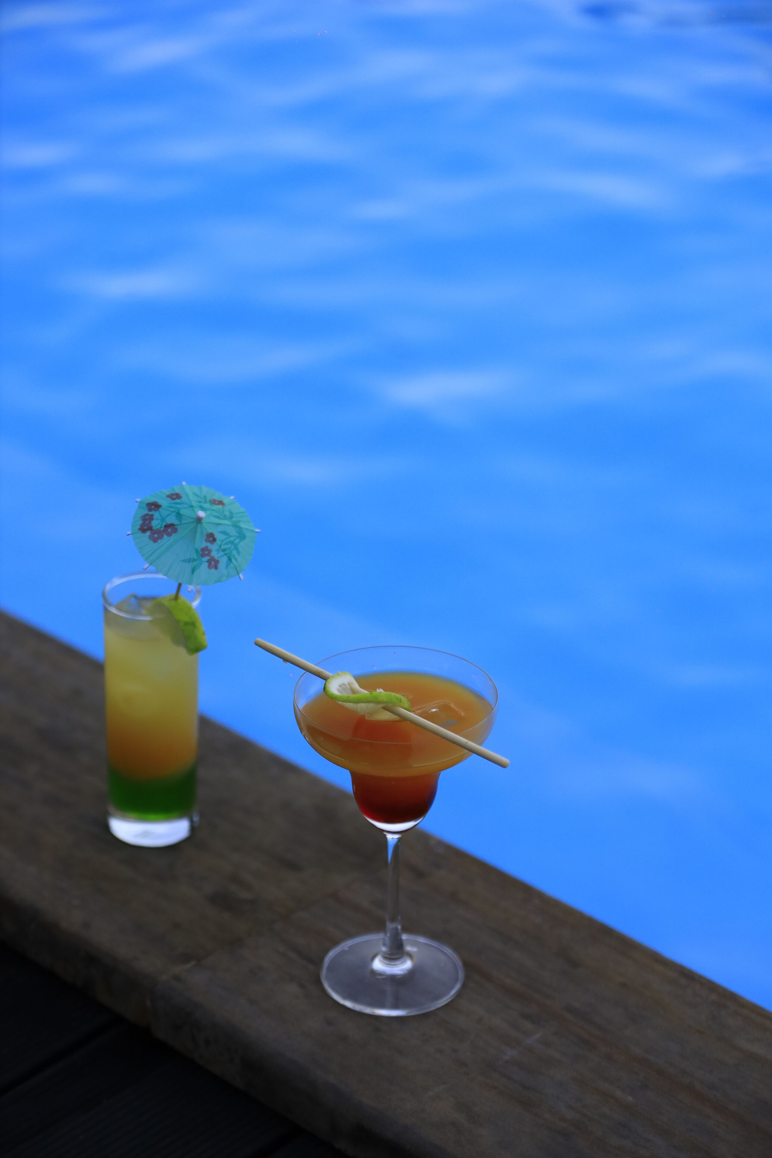 Two cocktails on a wooden ledge by a pool.