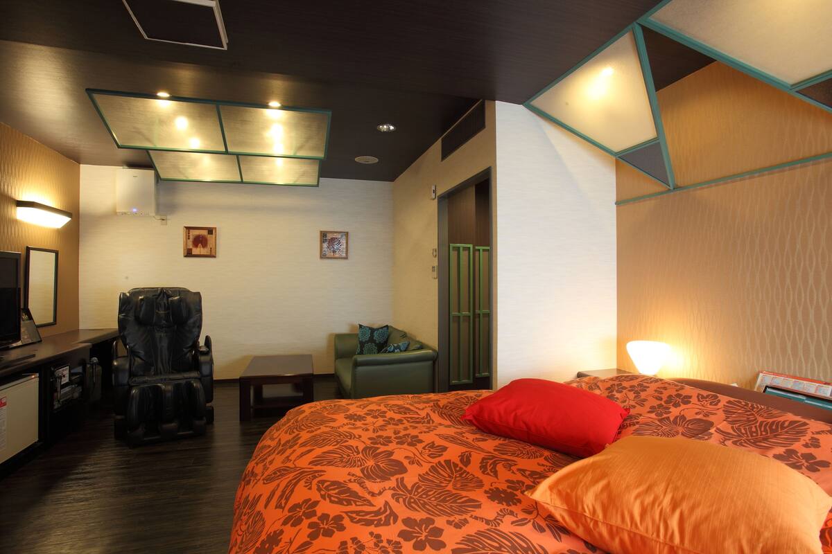 Quarto de hotel com cadeira de massagem, cama com roupa de cama floral, escrivaninha com computador e televisão.