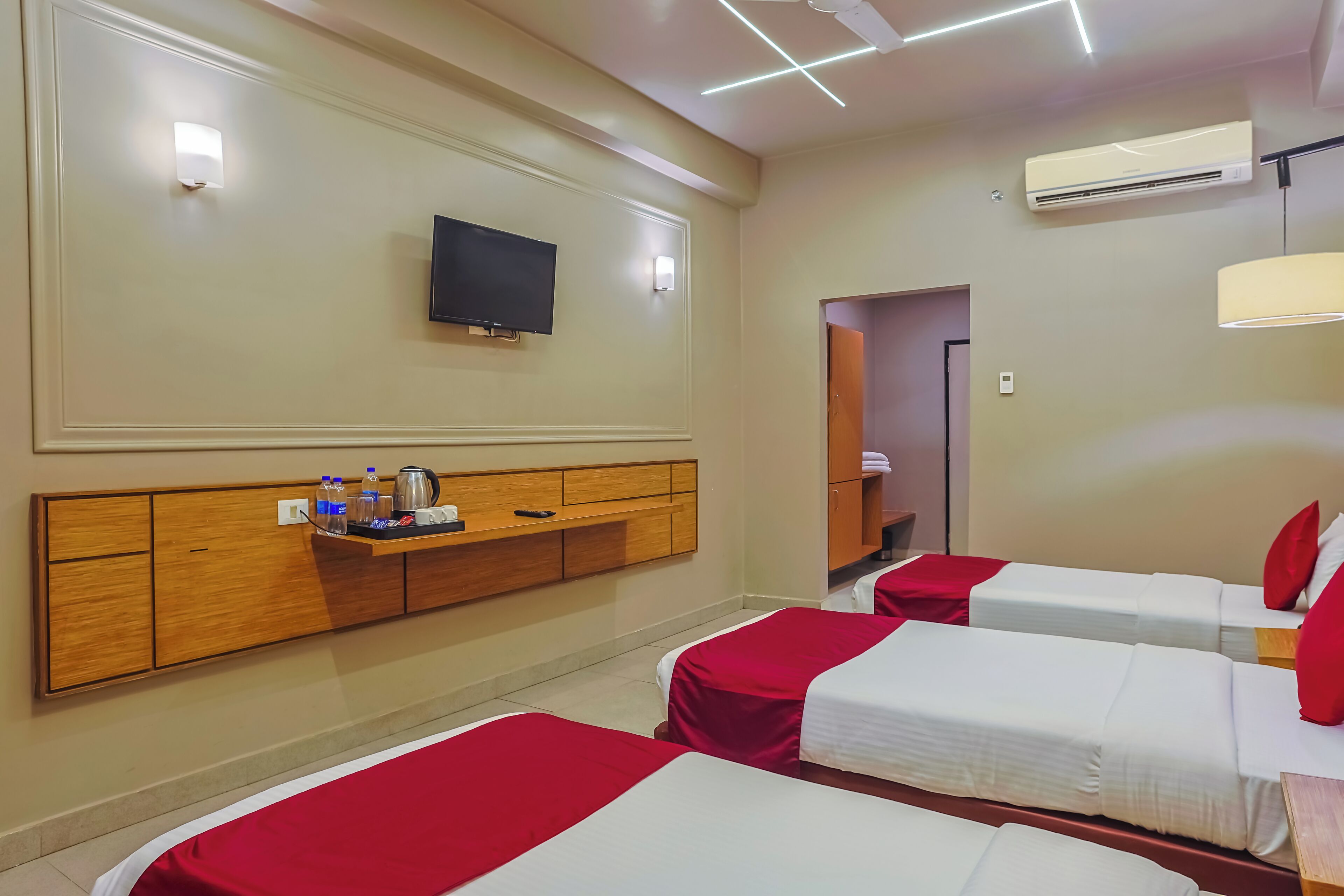 A hotel room with two beds, a TV mounted on the wall, a wooden headboard, a shelf with toiletries, and an air conditioning unit.