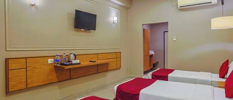 A hotel room with two beds, a TV mounted on the wall, a wooden headboard, a shelf with toiletries, and an air conditioning unit.