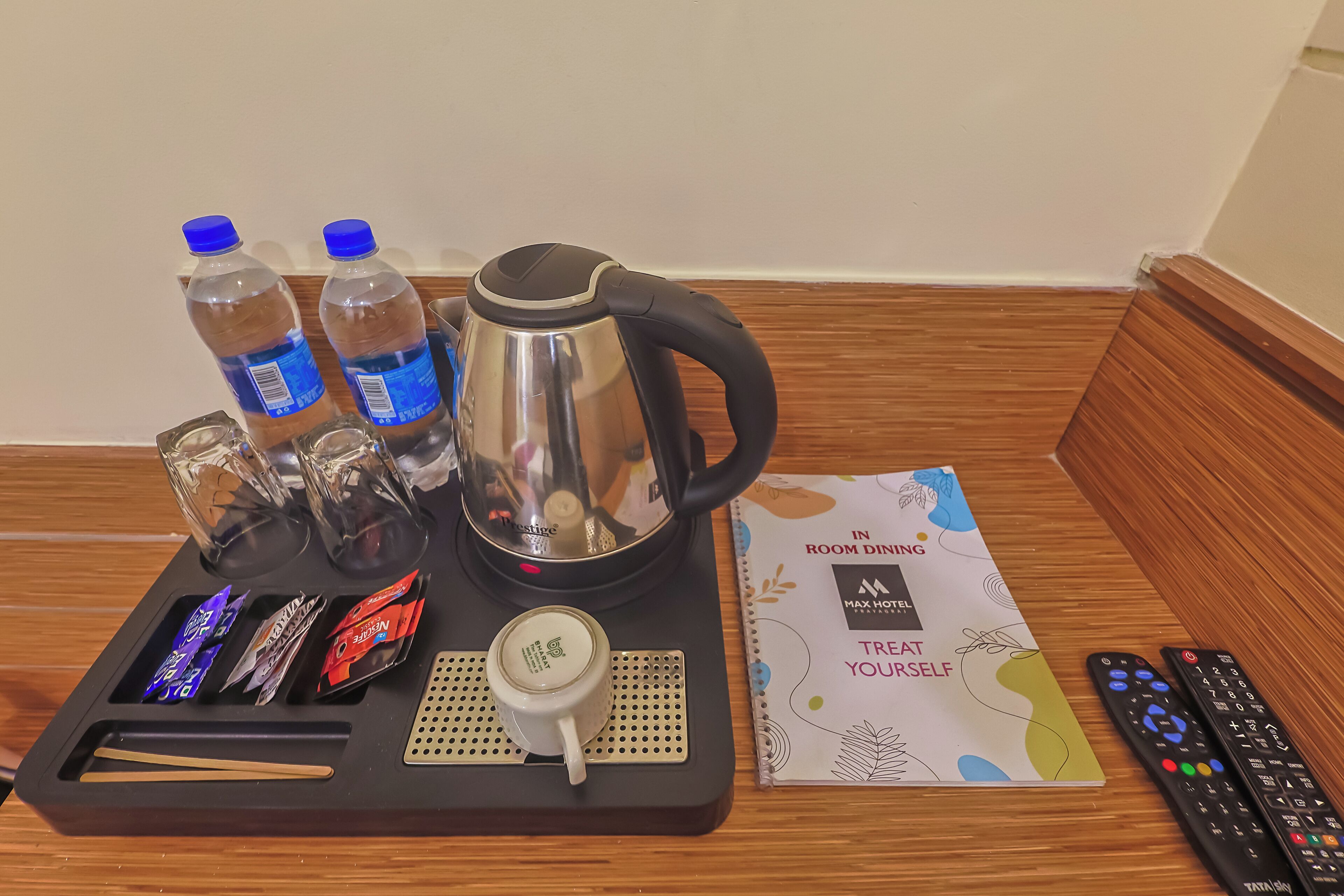 Een dienblad met een waterkoker, waterflessen, glazen, snacks, een mok en afstandsbedieningen op een houten vloer.