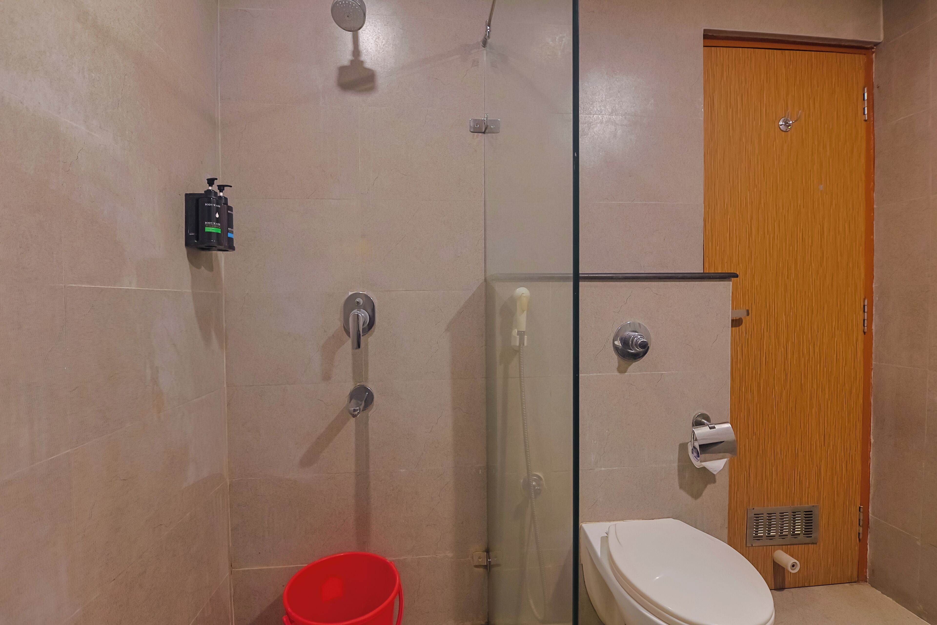 Une salle de bain avec une douche à parois de verre, des toilettes et un seau rouge.