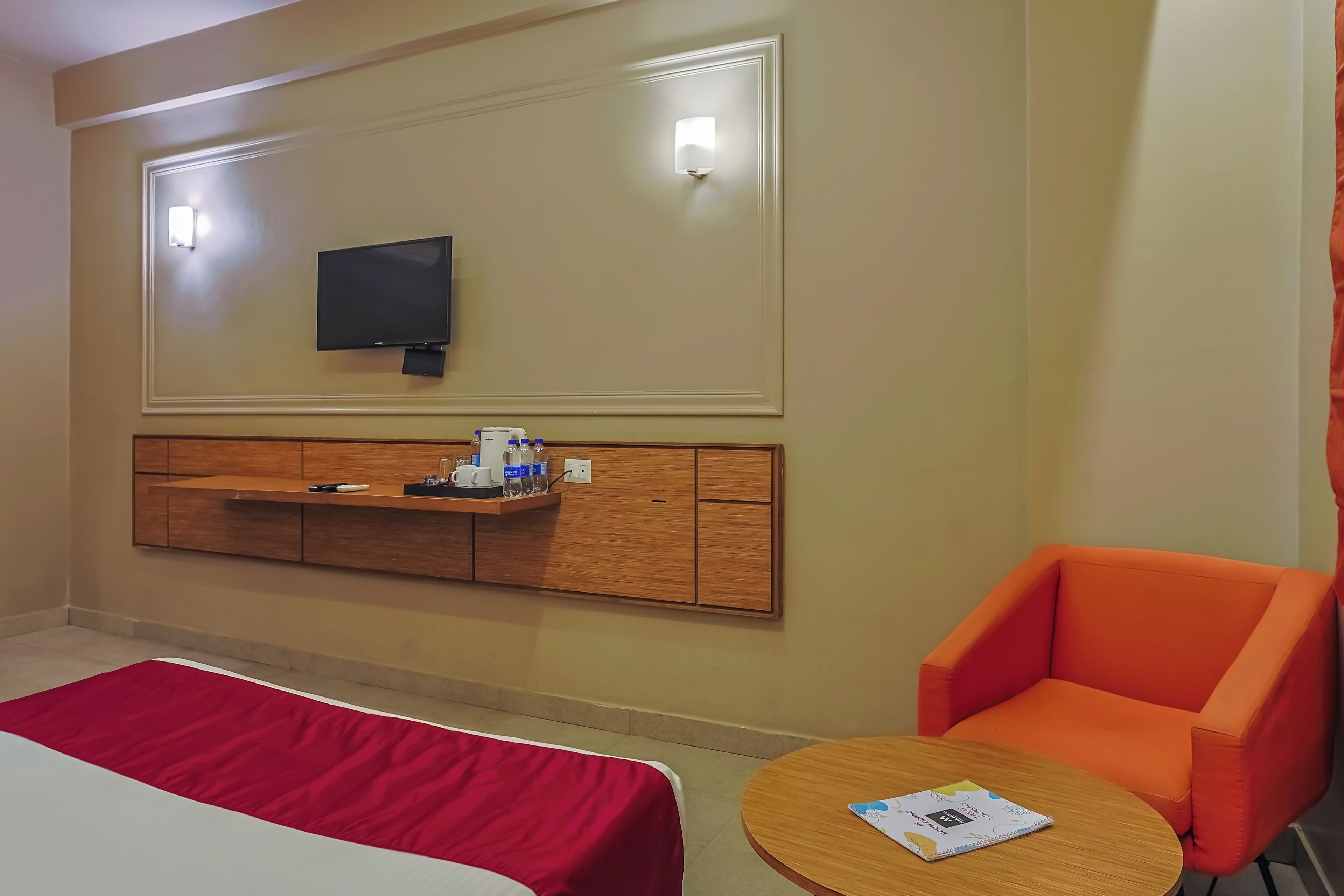 Una habitación de hotel con una cama, un edredón rojo, una mesita de noche de madera con un televisor, una pequeña mesa con un libro y una silla naranja.