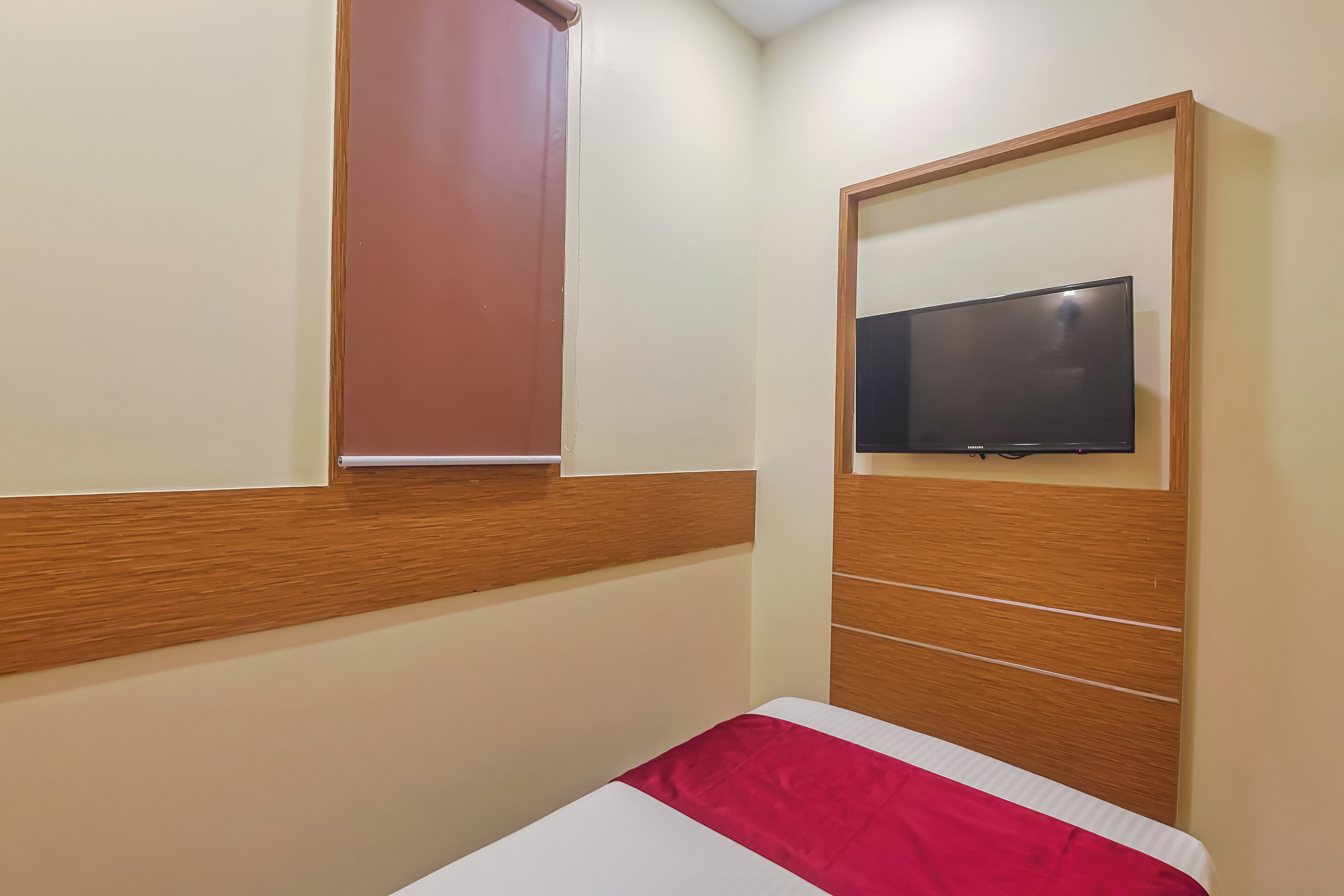 A hotel room with a bed, a television mounted on a wooden panel, and a window with a blind.